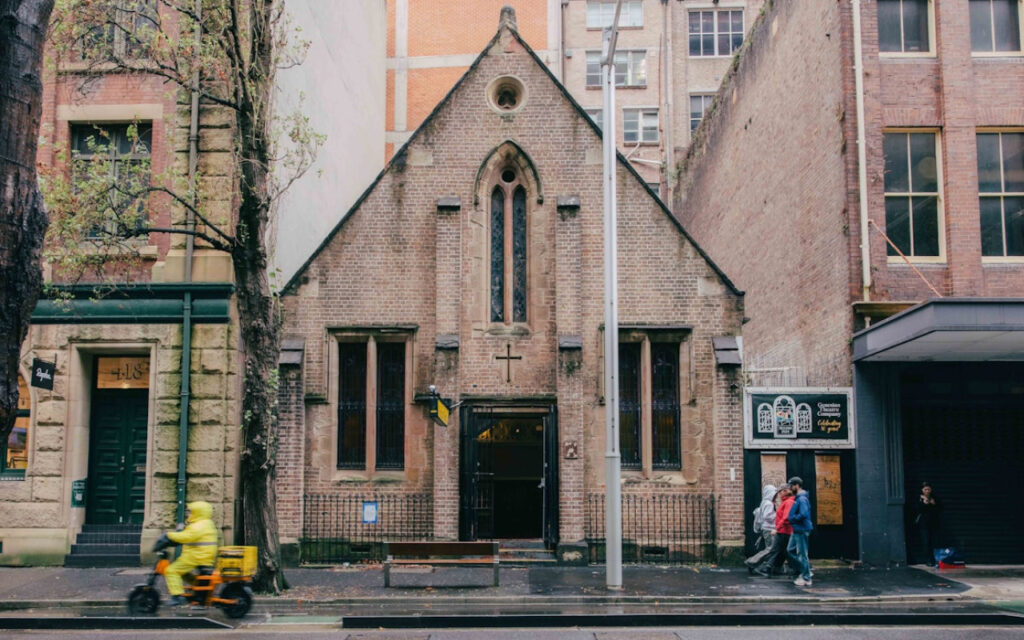 Small brick church with arched windows on a city street, between taller buildings; pedestrians pass by.