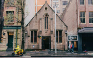 Small brick church with arched windows on a city street, between taller buildings; pedestrians pass by.