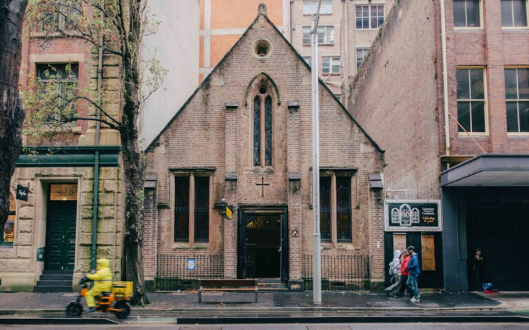 Small brick church with arched windows on a city street, between taller buildings; pedestrians pass by.
