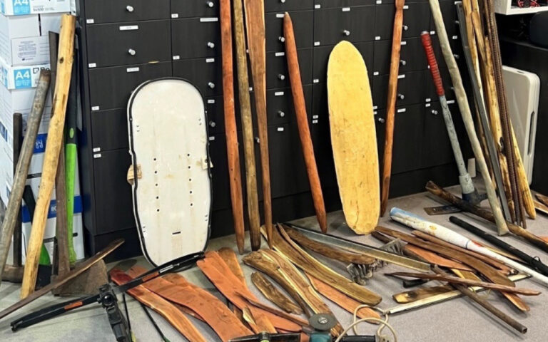 Collection of wooden oars and paddles leaning against dark storage lockers in a workshop. A white boat shield is propped nearby; scattered on the floor are more paddles and tools.