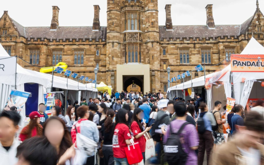 Crowd of people at an outdoor festival with white tents lining a historic brick building courtyard/entrance gate behind them.