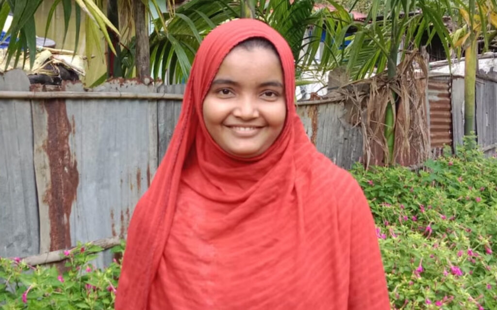 Smiling woman wearing a red hijab standing outdoors in a garden with a wooden fence behind her.