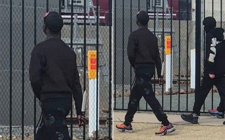 Three people in dark jackets and pants walk along a chain-link fence in an industrial area, wearing colorful sneakers and facing away from the camera.