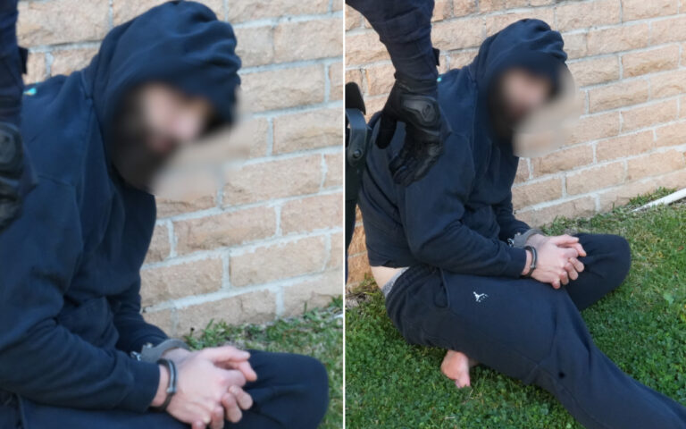 Person in a navy hoodie sits on grass against a brick wall with hands cuffed; an armed officer in gloves stands nearby, faces blurred.