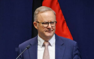 Man in navy suit and pink tie stands at a podium with a microphone, red flag in the background.