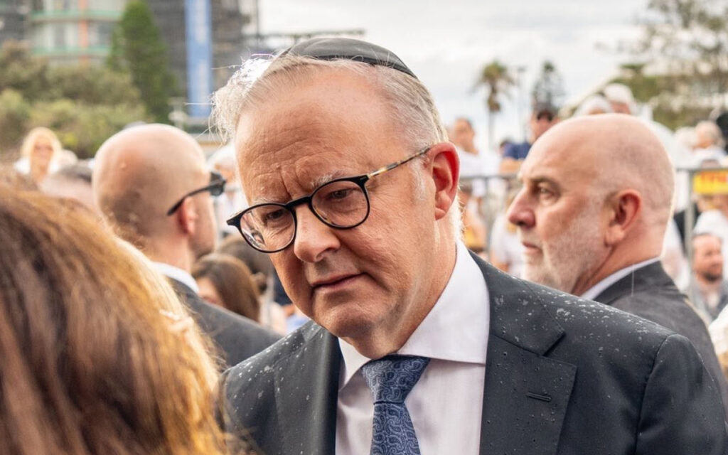 Older man wearing a kippah, glasses, and a dark suit talks with someone at a crowded outdoor event.