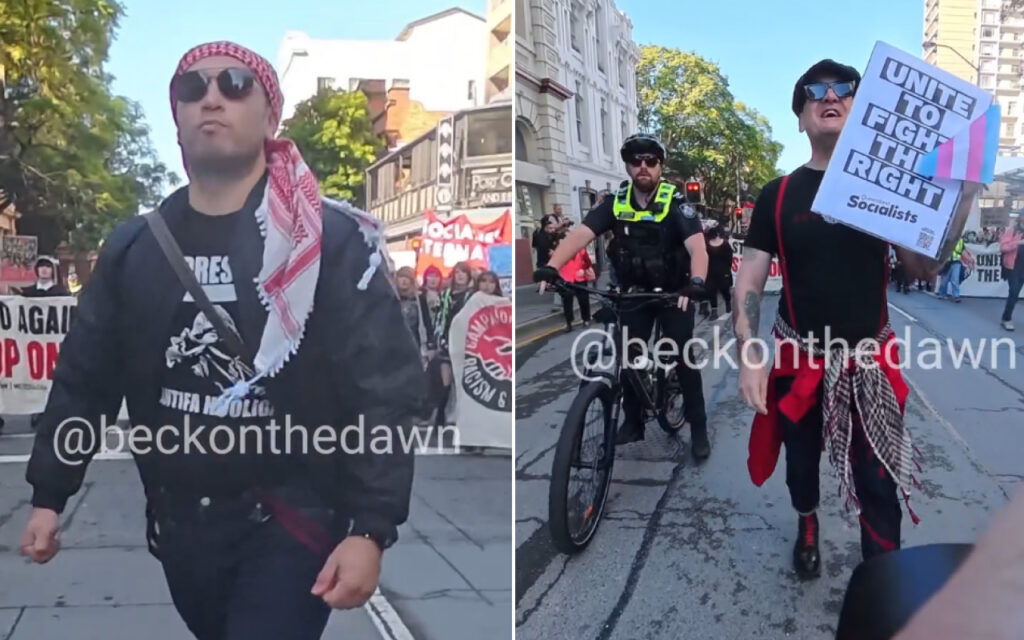 Two protesters walk toward the camera: left man wearing sunglasses and a red-and-white scarf, black jacket, and a graphic tee; crowd and signs in the background.