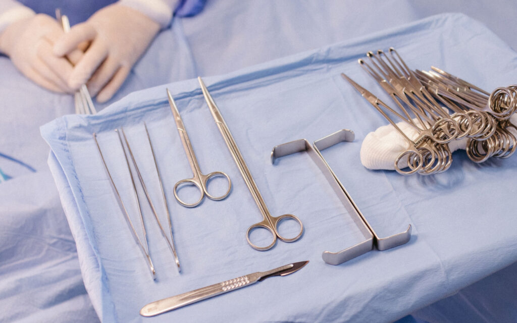 Surgical tray with stainless instruments on blue sterile drape: scissors, forceps, needle holders, scalpel, clamps, and a gloved hand in the background ready for procedure.