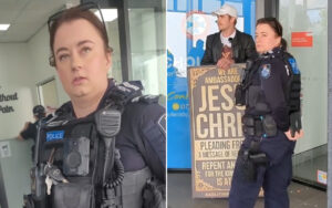 Two police officers in tactical gear indoors; one in the foreground with a radio and body camera, another beside a protest poster by a doorway.