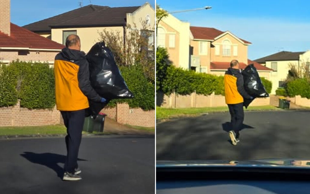 Man in an orange jacket carries a large black trash bag along a residential street, back to camera during daytime walk-up cleanup.