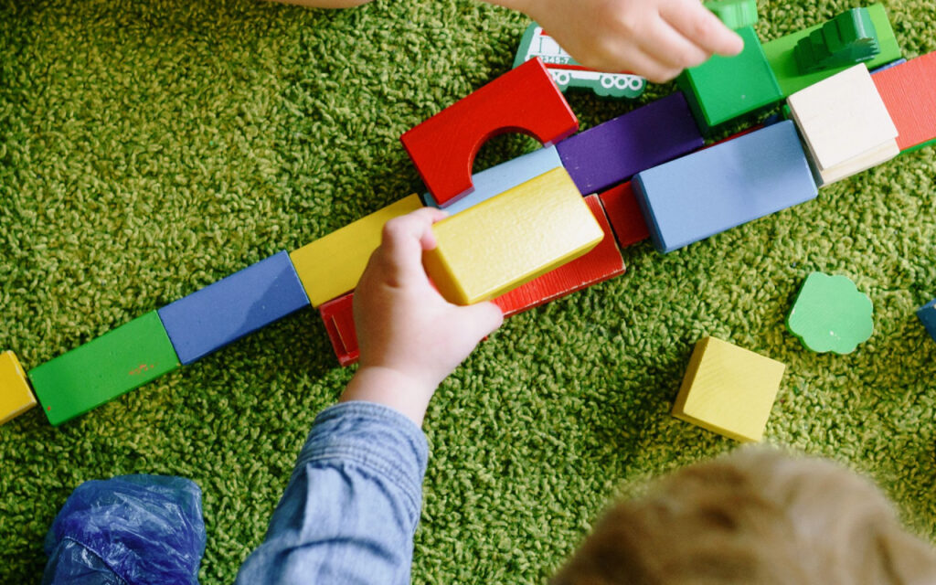Child's hands placing a yellow wooden block onto a line of colorful blocks on a green carpeted floor.