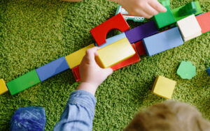 Child's hands placing a yellow wooden block onto a line of colorful blocks on a green carpeted floor.
