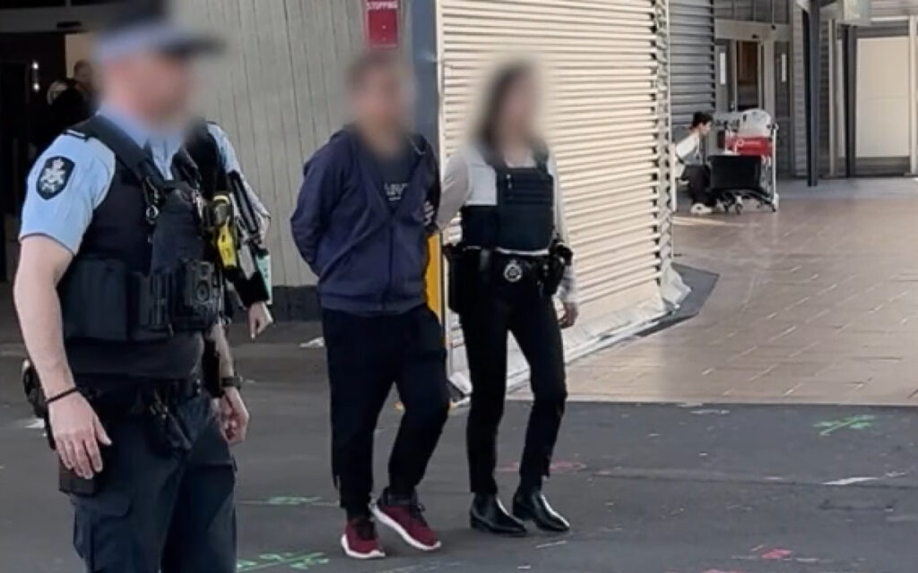 Blurred civilians escorted by a police officer down a city sidewalk.