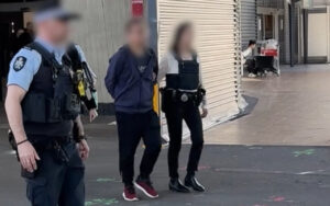 Blurred civilians escorted by a police officer down a city sidewalk.