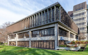 Concrete, two-story office building with large windows and a brown vertical slat facade on the upper level, set on a grassy plaza.