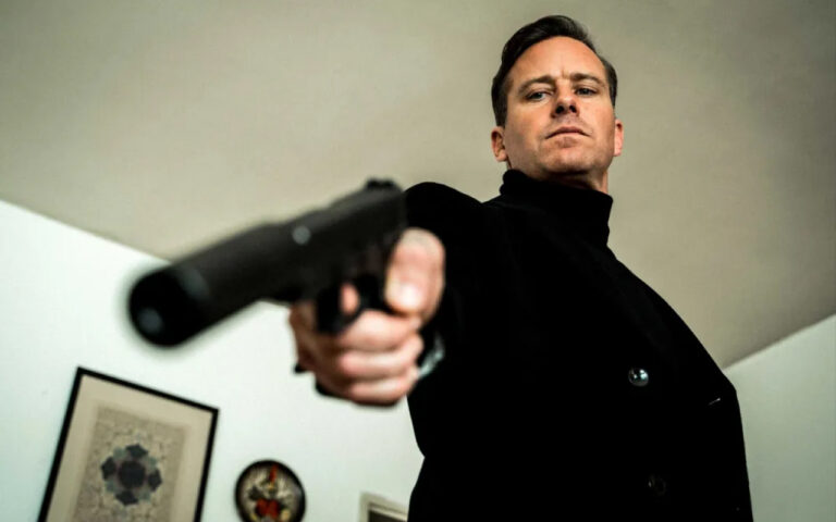 Man in a black coat points a handgun toward the camera from a low, dramatic angle in an indoor setting.