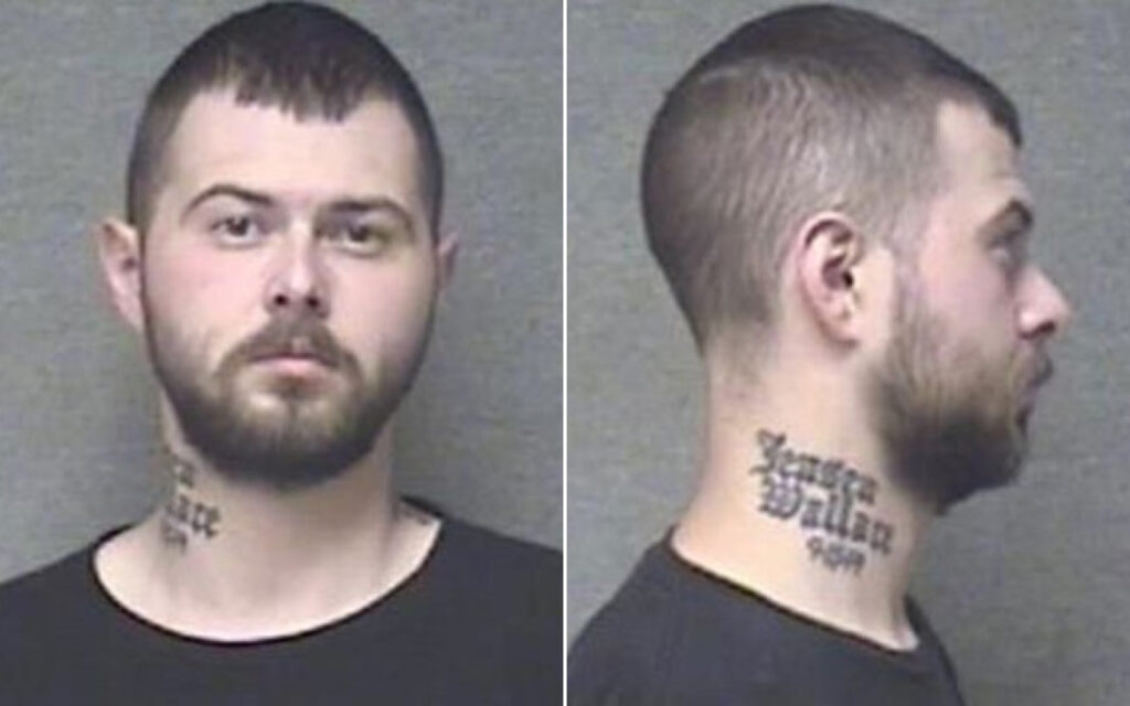 Mugshot of a bearded man with short dark hair, front view, neck tattoo visible on left side; black shirt, neutral background