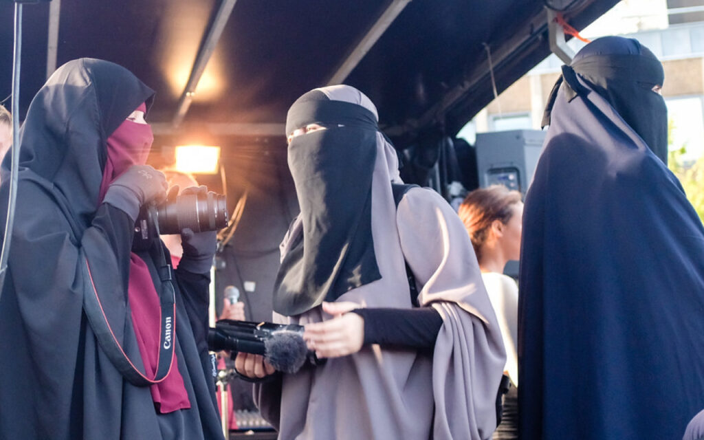 Women wearing abayas and niqabs filming with professional cameras on a set or street scene under a canopy.
