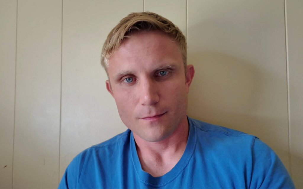 Portrait of a man with short blond hair and blue eyes, wearing a bright blue t-shirt, looking at the camera against a pale wall backdrop.