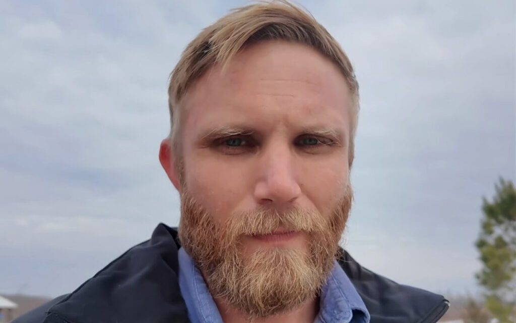 Man with blond hair and a full reddish-blonde beard, wearing a blue collared shirt and dark jacket, outdoors on a cloudy day.