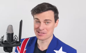 Man in a blue blazer with a Union Jack pattern on the sleeve speaks into a studio microphone in front of a plain background.