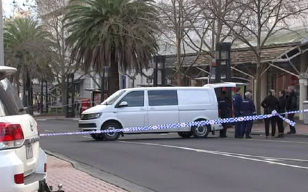 White van stopped on a street behind police tape, with several people gathered nearby as investigators work the scene.