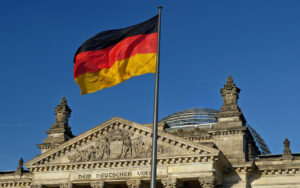 German flag waving in front of the Reichstag building, with a sculpted triangular pediment and glass dome in clear blue sky