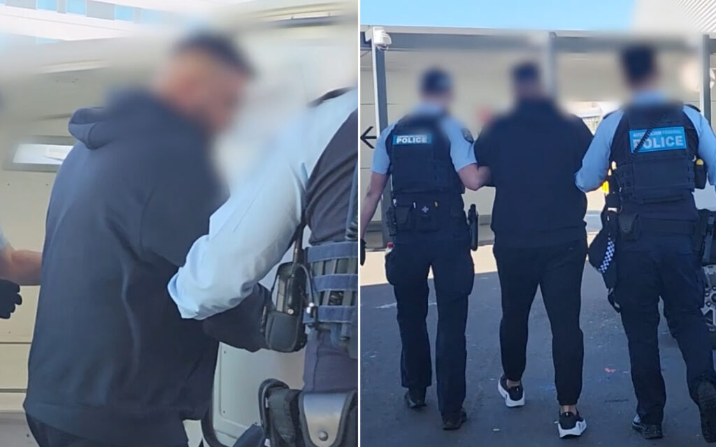Two police officers escort a man toward a vehicle; one officer holds his arm as another stands nearby in uniform.
