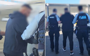 Two police officers escort a man toward a vehicle; one officer holds his arm as another stands nearby in uniform.