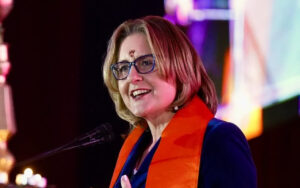 Smiling blonde woman with glasses speaking into a microphone on a stage, wearing an orange scarf over a blue blazer.