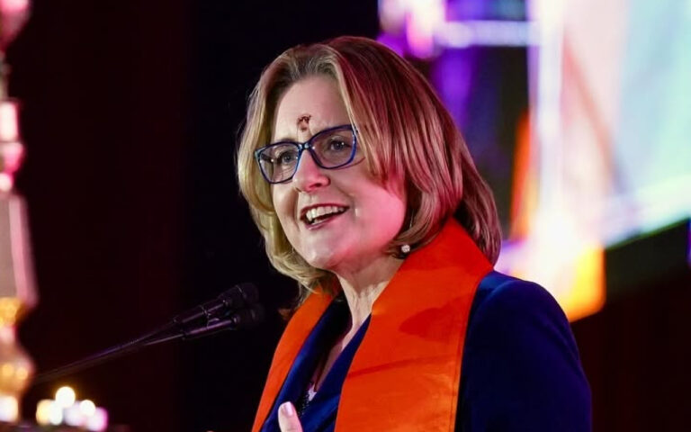 Smiling blonde woman with glasses speaking into a microphone on a stage, wearing an orange scarf over a blue blazer.
