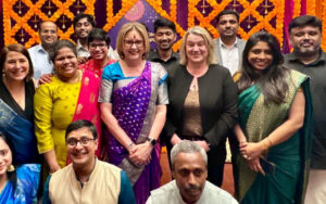 Group of colleagues and friends posing together at a festive event with orange marigold decorations in the background, smiling.