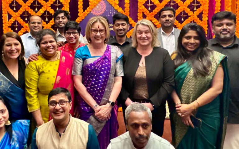 Group of colleagues and friends posing together at a festive event with orange marigold decorations in the background, smiling.