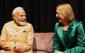 Two adults sit on a pink sofa, smiling and talking on a stage with a dark backdrop.