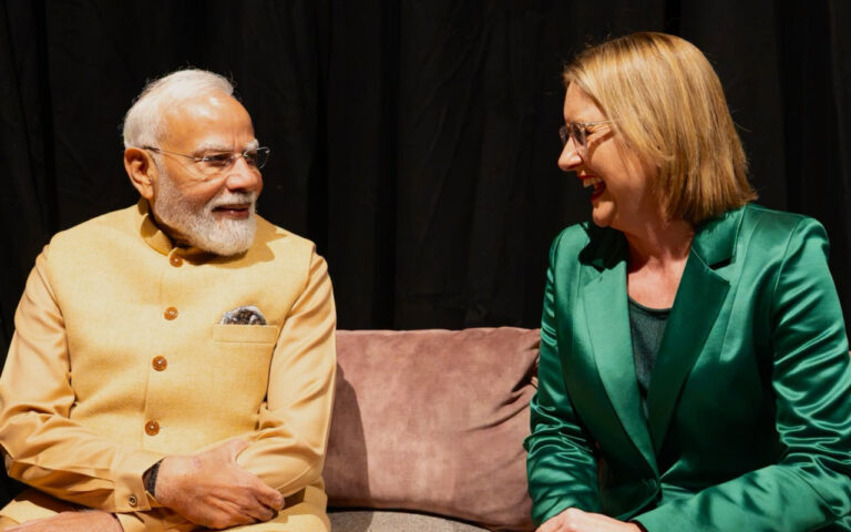 Two adults sit on a pink sofa, smiling and talking on a stage with a dark backdrop.