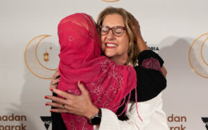 Two women share a warm hug on a stepped red-carpet backdrop, one wearing a pink embroidered headscarf and the other smiling with glasses.