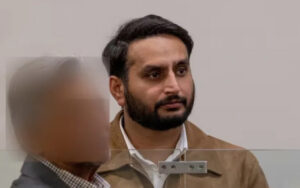 Bearded man in a brown jacket with white shirt, looking to the right, in a meeting setting behind a glass divider with a blurred foreground figure.