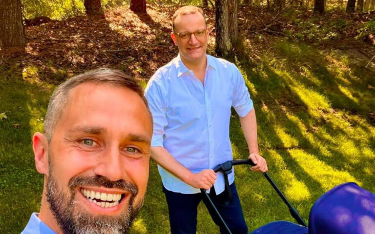 Two men outdoors on a sunny day in a park; the bearded man in the foreground smiles at the camera while the other man pushes a blue stroller in the background.