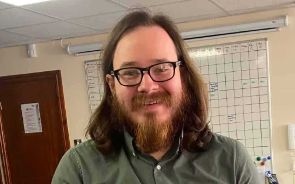 Man with shoulder-length brown hair, a full beard, and glasses smiles at the camera in an office wearing a green button-down shirt, with a whiteboard calendar in the background.