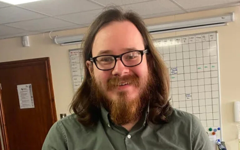 Man with shoulder-length brown hair, a full beard, and glasses smiles at the camera in an office wearing a green button-down shirt, with a whiteboard calendar in the background.