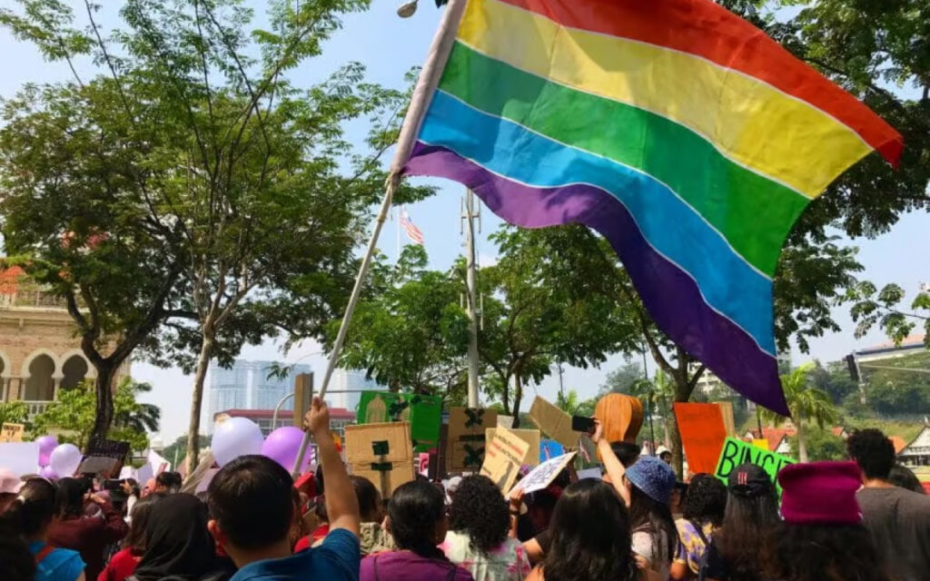 Crowd of protesters outdoors holding signs, with a large rainbow Pride flag waving overhead.