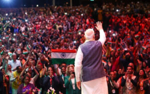 Speaker on stage waving to a large cheering crowd holding Indian flags at a political rally at night.