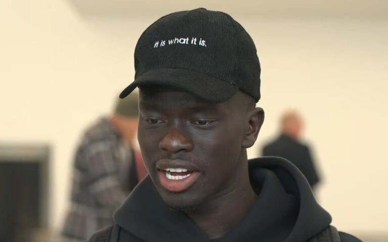 Young Black man wearing a black cap with the text 'hi is what it is,' speaking softly with a blurred background behind him, wearing a hoodie.