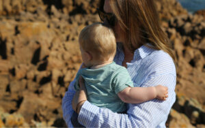 Woman in a blue striped shirt holding a baby outdoors, with a rocky cliff background and sunny weather.