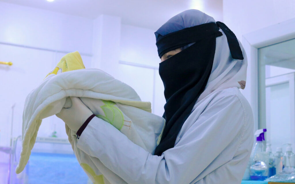 Person wearing a hijab and niqab folds a pale baby blanket in a clinical hospital room.