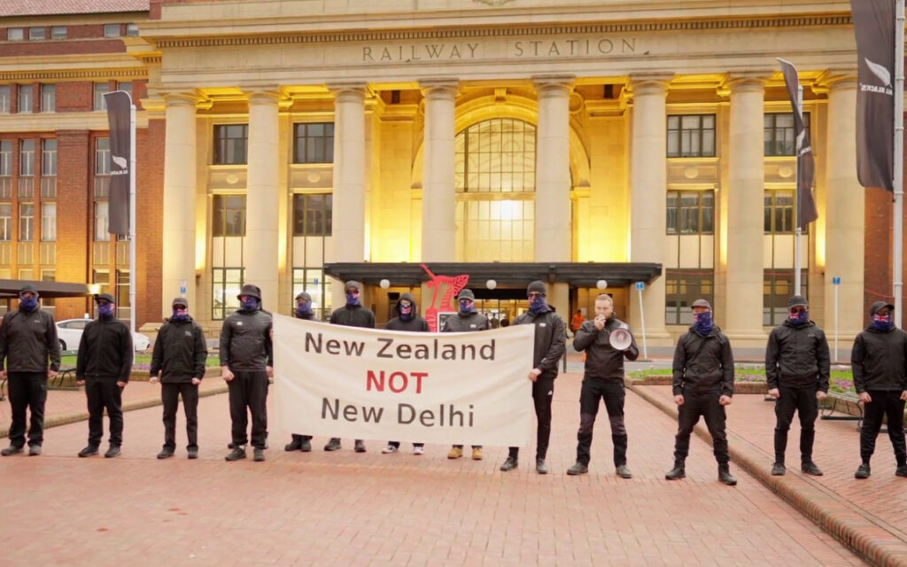Masked protesters in black clothing stand before a grand railway station, holding a banner that reads 'New Zealand NOT New Delhi'.