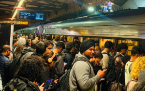 Crowded subway platform with a long silver train at the doors; passengers with backpacks and phones wait together.