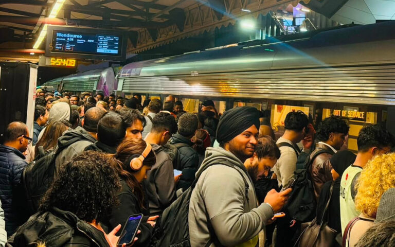 Crowded subway platform with a long silver train at the doors; passengers with backpacks and phones wait together.