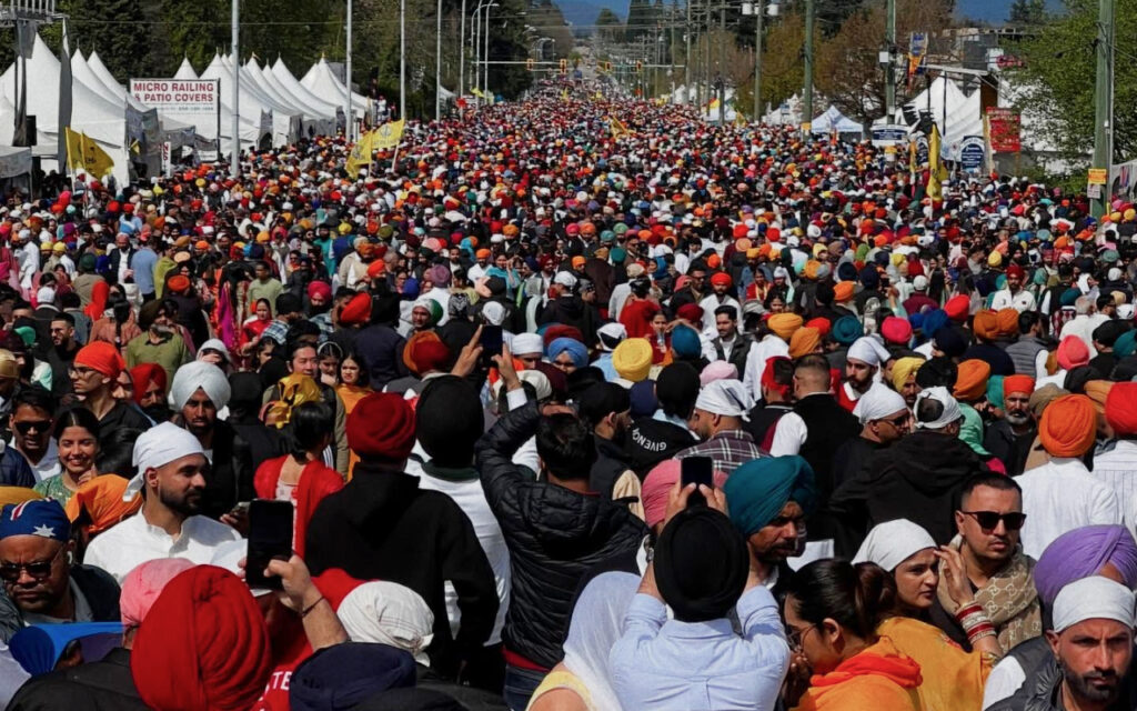 Massive crowd of people wearing colorful turbans fills a street festival with white tents lining both sides and signs in the distance.