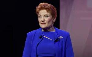 Speaker with short red hair in a royal blue blazer giving a talk on stage, with lapel mics visible.